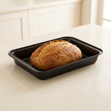 A round artisanal loaf of bread sits inside a rectangular black baking pan on a light cream countertop, with a softly blurred kitchen background.