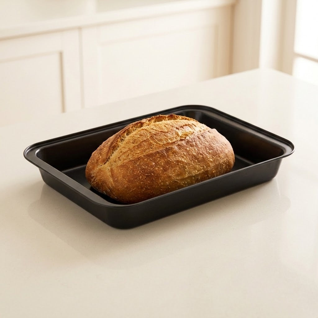 A round artisanal loaf of bread sits inside a rectangular black baking pan on a light cream countertop, with a softly blurred kitchen background.