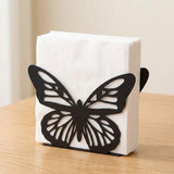 A black butterfly-shaped paper towel or napkin holder holding a stack of white paper towels, set on a light wood tabletop against a soft beige background.