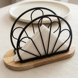 Black metal napkin holder with a wooden base, holding white folded napkins, set on a light beige table with a blurred plate in the background.