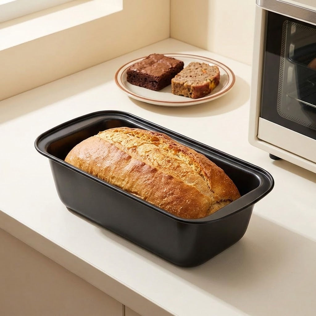 A black rectangular loaf pan containing a freshly baked loaf of bread, set on a kitchen counter with a plate of brownies and a microwave in the background.