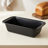 A black rectangular loaf pan sits on a light beige countertop, with a golden loaf of bread on a wooden board blurred in the background.