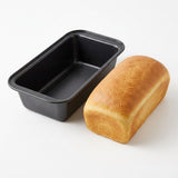 A black rectangular loaf pan shown empty next to a golden-brown baked loaf of bread on a clean white background.