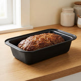 A black rectangular loaf pan containing a baked meatloaf, placed on a wooden countertop in a kitchen setting with cream-colored canisters and a window in the background.