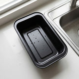 A black rectangular non-stick baking pan with a raised center ridge, resting on a white kitchen countertop next to a stainless steel sink.