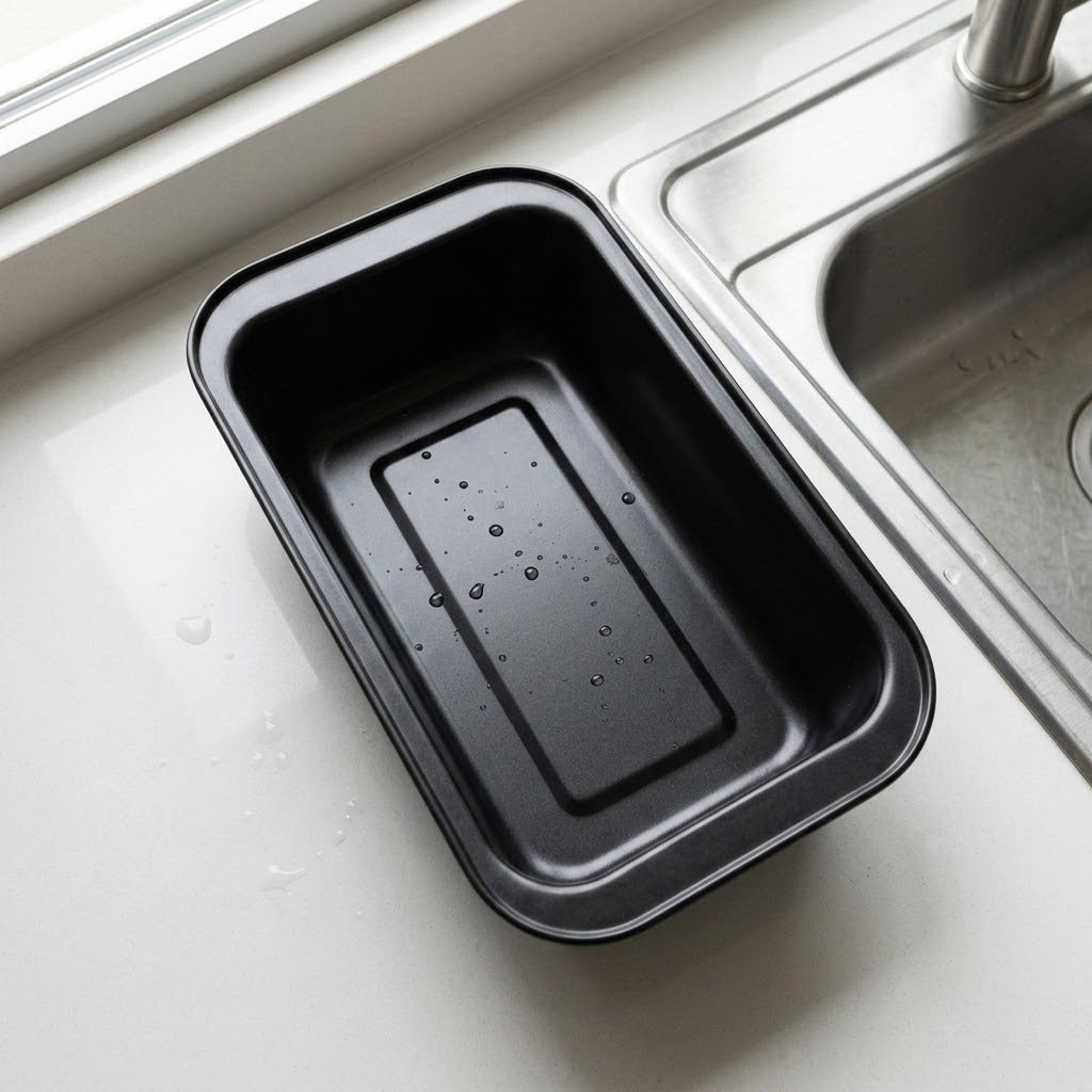 A black rectangular non-stick baking pan with a raised center ridge, resting on a white kitchen countertop next to a stainless steel sink.