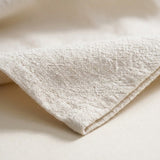 A close-up of a folded cream-colored textured fabric, likely cotton or linen, showing its woven detail and soft shadows.