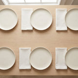 Six minimalist off-white ceramic plates arranged in two rows on a light wood table, alternating with folded white napkins.