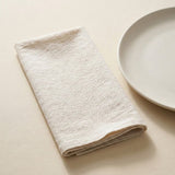 A folded cream-colored textured cloth napkin or tea towel placed on a light beige surface next to a white ceramic plate.