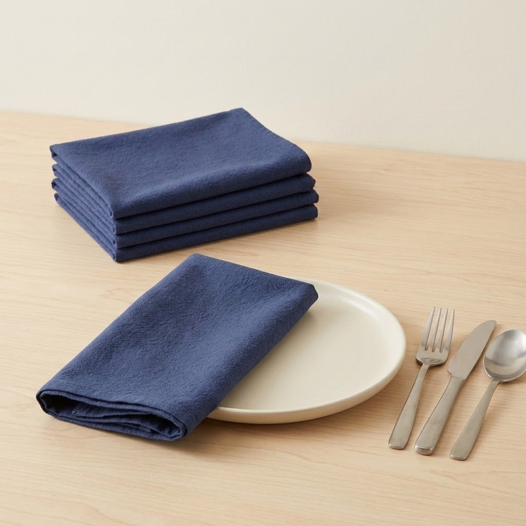 A set of folded navy blue linen napkins on a light wood table, with one napkin displayed on a white plate and three silver utensils beside it.