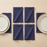 Six navy blue folded cloth napkins arranged on a light wood tabletop with white plates partially visible at the left and right edges.