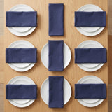 Nine navy blue napkins are neatly folded and arranged in a grid pattern over white plates on a light wood tabletop.