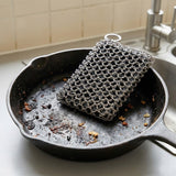 A silver metal chainmail scrubber resting inside a dark cast iron skillet on a kitchen counter near a sink.
