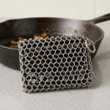 A silver metal mesh scrubber block with a hanging ring, resting in front of a dark cast iron skillet on a light gray countertop.
