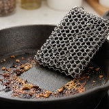 A rectangular steel chainmail scrubber resting inside a dark cast iron skillet with burnt food residue, demonstrating its use for cleaning.