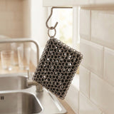 A stainless steel wire mesh sponge scrubber hangs from a metal hook mounted on a white kitchen cabinet, with a sink and tiled wall in the background.