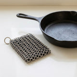 A square metal chainmail scrubber with a key ring, shown next to a black cast iron skillet on a light cream-colored surface.