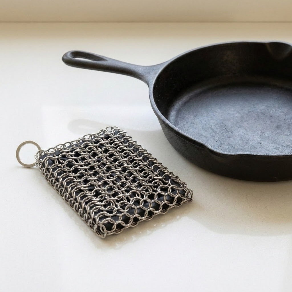 A square metal chainmail scrubber with a key ring, shown next to a black cast iron skillet on a light cream-colored surface.