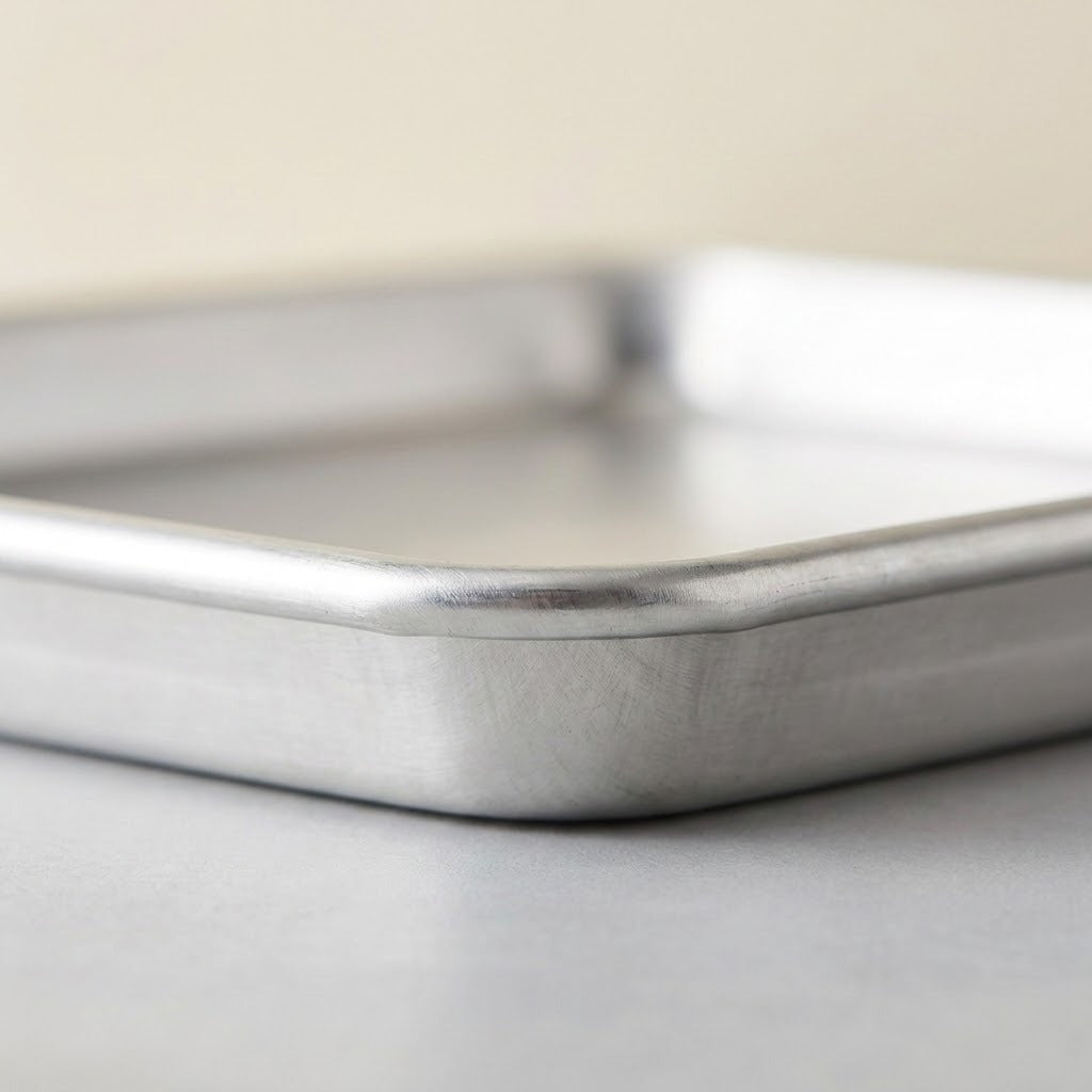 A close-up shot of a silver metal baking pan showing its thick rim and interior corner, photographed on a neutral light gray surface with a soft beige background.