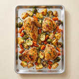 A top-down view of a roasted chicken and vegetable sheet pan meal on a silver metal baking tray, set against a solid beige background.