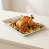 A rectangular metal roasting pan containing a roasted whole chicken with carrots, potatoes, and Brussels sprouts, photographed on a light cream countertop in a bright kitchen setting.