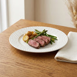 A white textured ceramic plate holding sliced steak, green asparagus, and roasted potatoes, set on a wooden table with a folded cream napkin nearby.