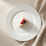 A white ceramic plate with a textured, dimpled border, styled on a beige fabric background with a slice of cheesecake topped with raspberries and blueberries in the center.