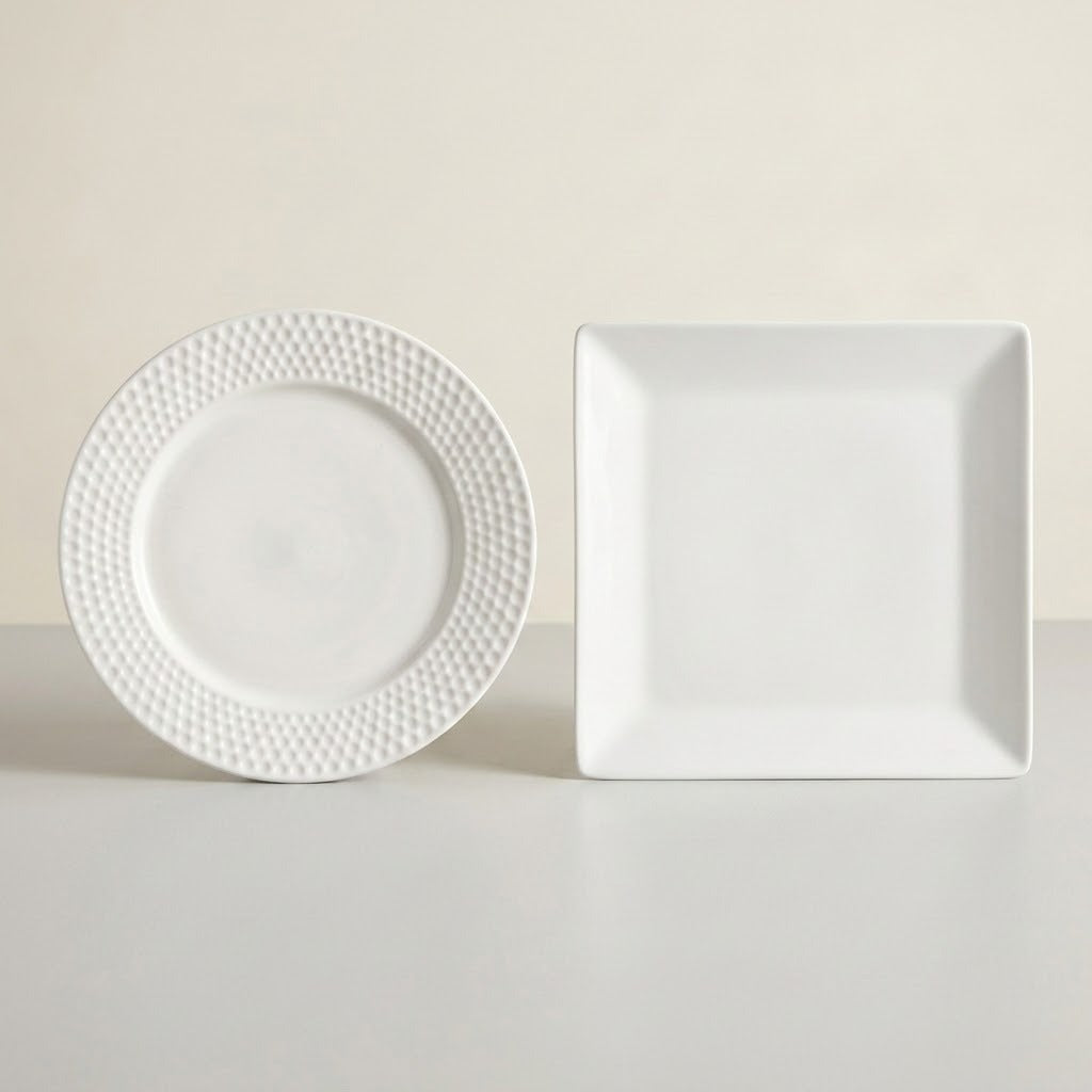 A white ceramic dinner plate with a textured beaded rim and a matching square white plate, displayed on a light gray surface against a warm beige background.