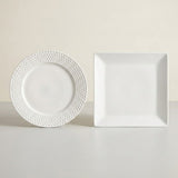 A white ceramic dinner plate with a textured beaded rim and a matching square white plate, displayed on a light gray surface against a warm beige background.