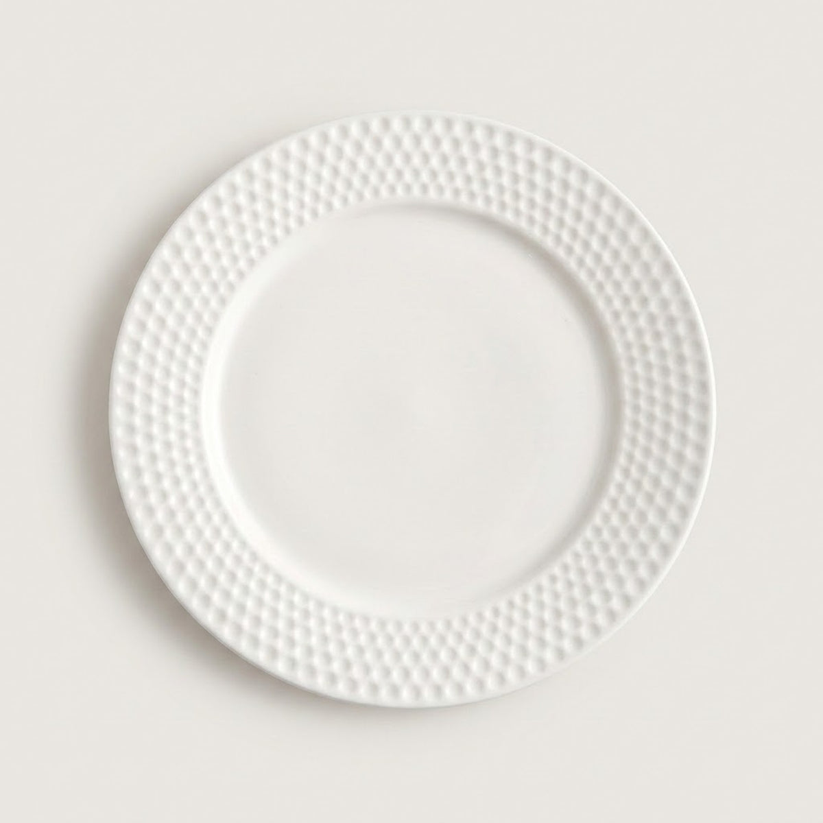 A white ceramic dinner plate with a textured, dimpled pattern around the rim, shown from a top-down view on a light beige background.