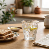 A textured clear glass tumbler with a lemon slice, filled with a bubbly drink, set on a wooden table next to a plate of toast in a cozy kitchen setting.