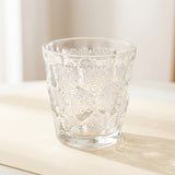 A clear textured glass tumbler with a diamond-patterned design, sitting on a light beige surface with a soft-focus background.