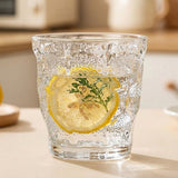 A clear textured glass tumbler with a lemon slice and green herb garnish inside, set on a light tabletop with a softly blurred kitchen background.
