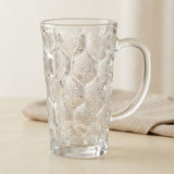A clear glass mug with a textured honeycomb pattern and a curved handle, shown on a light beige tabletop with a soft neutral background.