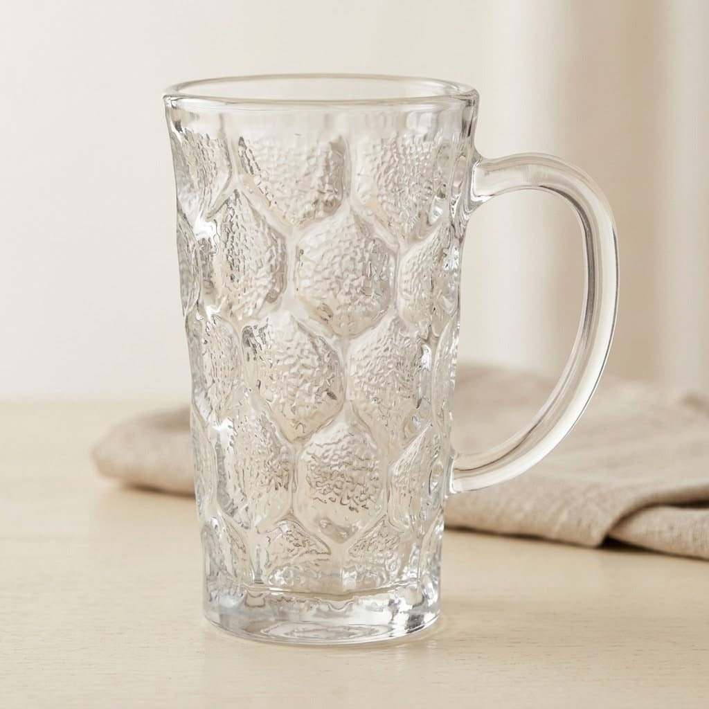 A clear glass mug with a textured honeycomb pattern and a curved handle, shown on a light beige tabletop with a soft neutral background.