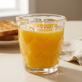 A clear textured glass tumbler filled with orange juice, placed on a light tabletop with toast and a cloth in the background.