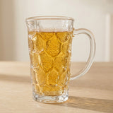 A clear textured glass mug filled with an amber beverage, set on a light wood surface against a soft beige background.