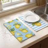 Two kitchen towels or placemats on a countertop near a sink: one with a lemon and leaf print on light blue, and one with light blue and white stripes and a white plate on top. A spilled drink is visible on the left side.