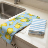 A kitchen towel set displayed on a white countertop next to a sink. One towel is light blue with a yellow lemon and green leaf pattern, and the other is folded in a light blue and white striped fabric.