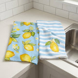Two kitchen towels with a lemon print and striped pattern resting on a white countertop next to a stainless steel sink.