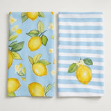 Two cotton kitchen towels with a lemon theme, displayed side-by-side against a light neutral background. The towel on the left features a light blue background with a yellow lemon and green leaf pattern, while the towel on the right features light blue and white horizontal stripes with a single yellow lemon illustration.
