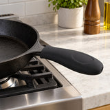 Black silicone pot handle cover shields a skillet handle beside a stovetop in a bright kitchen.