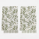 Two white kitchen towels with a green leaf pattern and green plaid borders, displayed side-by-side on a white background.