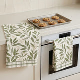 Two kitchen towels with a green leaf pattern hanging on a white oven handle and counter, with a baking tray of cookies on the counter in a bright kitchen setting.