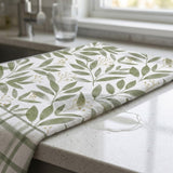 A folded kitchen towel with a green leaf pattern and striped border rests on a light speckled countertop near a sink and window.