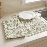 A set of kitchen placemats with a green leaf pattern on a cream background, styled on a kitchen counter with white plates and a glass on a sink.
