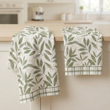 Two kitchen towels with a green leaf pattern and plaid borders hanging over a light wood countertop in a softly blurred kitchen setting.