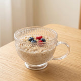 A clear textured glass mug filled with oatmeal and topped with blueberries and raspberries, set on a light wood table.