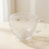 A clear glass teacup with an embossed floral and geometric pattern, featuring a sturdy handle and a small pedestal base, set on a light cream surface with a soft shadow.