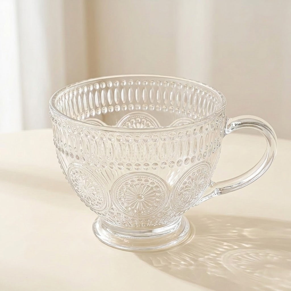 A clear glass teacup with an embossed floral and geometric pattern, featuring a sturdy handle and a small pedestal base, set on a light cream surface with a soft shadow.